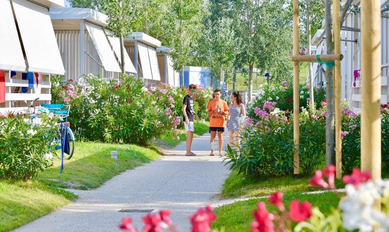Tre personer går på en sti omgivet af blomster og huse.
