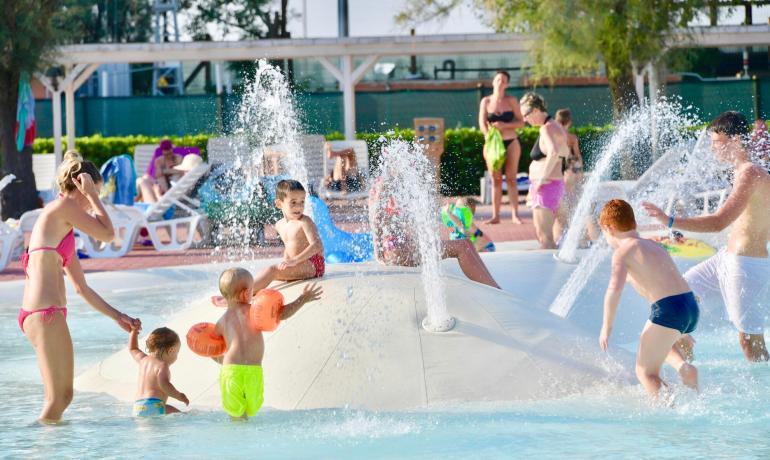 Children play in the pool with water sprays and adults in the background.