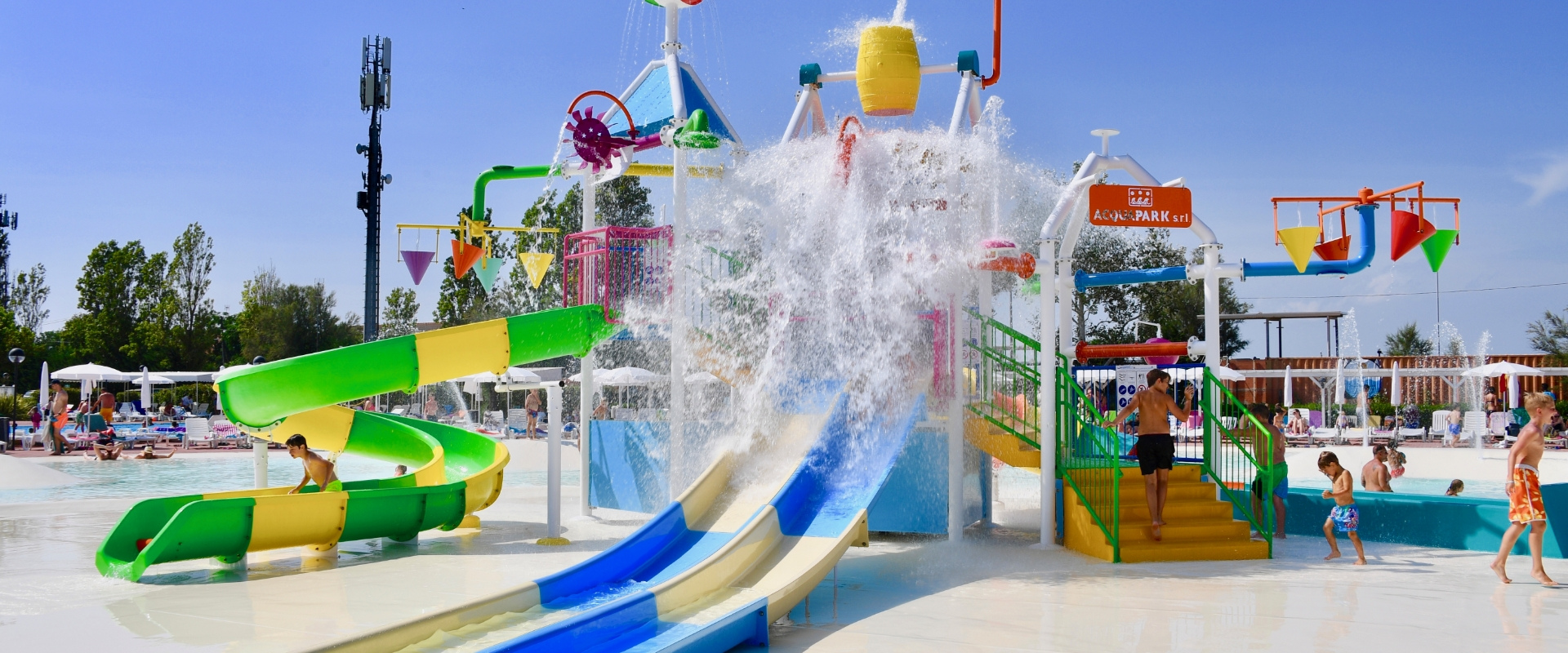 Bambini giocano in piscina con spruzzi d'acqua, sotto un cielo azzurro.
