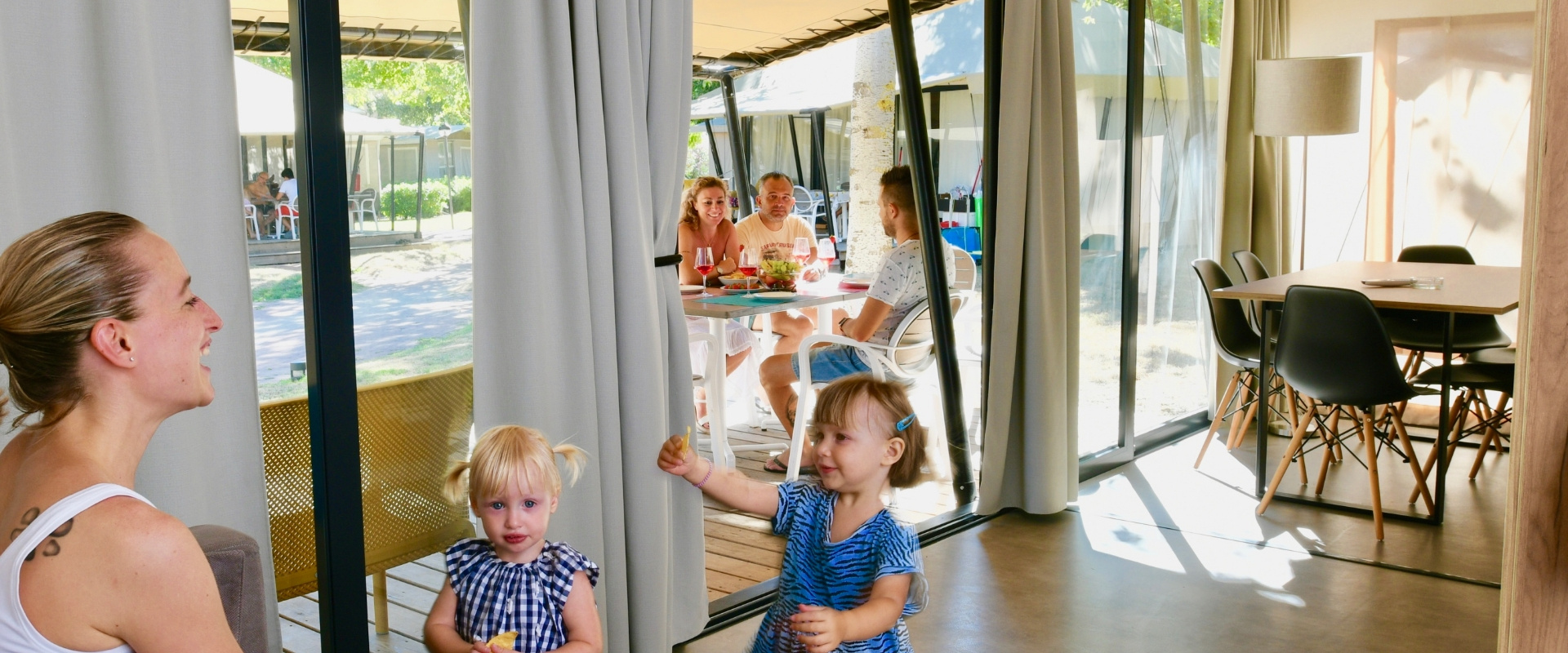 Famiglia che pranza all'aperto vicino a una tenda, bambini giocano all'interno.