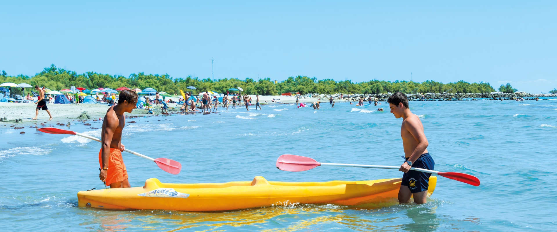 Due persone portano un kayak giallo in mare su una spiaggia affollata.