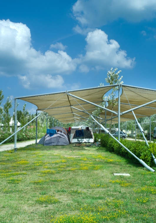 Tenda da campeggio sotto una struttura ombreggiante in un'area verde con cielo sereno.