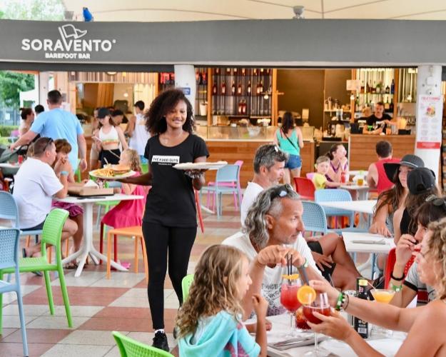 Bar affollato all'aperto con cameriera che serve clienti sorridenti, atmosfera vivace e colorata.