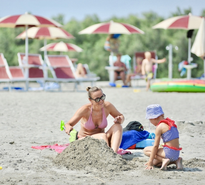 Donna e bambina giocano con la sabbia in spiaggia, sotto ombrelloni rossi e bianchi.