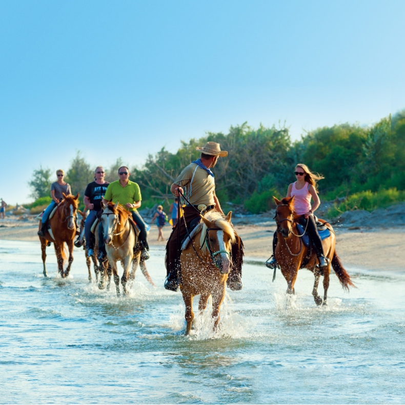 Persone a cavallo lungo la spiaggia, attraversando l'acqua sotto un cielo sereno.