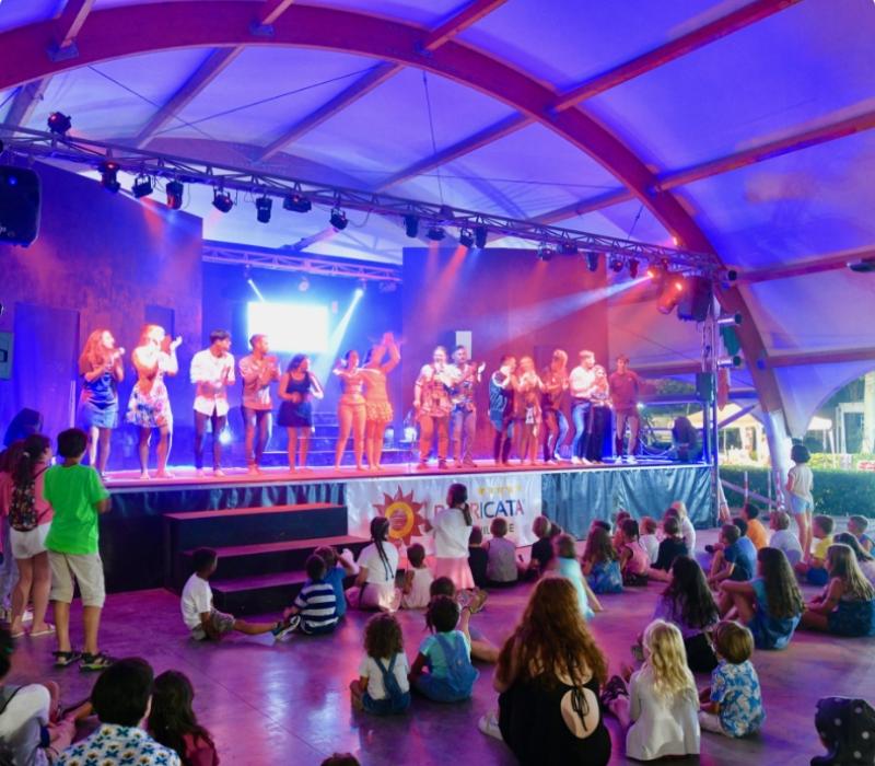 Spettacolo teatrale con bambini seduti ad assistere sotto un tendone illuminato.