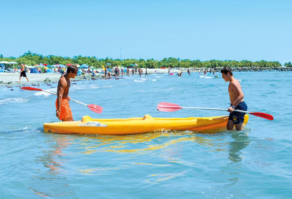 Due persone con un kayak giallo in mare, spiaggia affollata sullo sfondo.