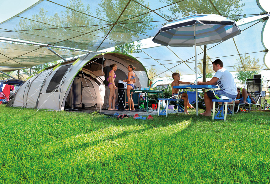 Famiglia in campeggio con tenda, tavolo da picnic e ombrellone su prato verde.